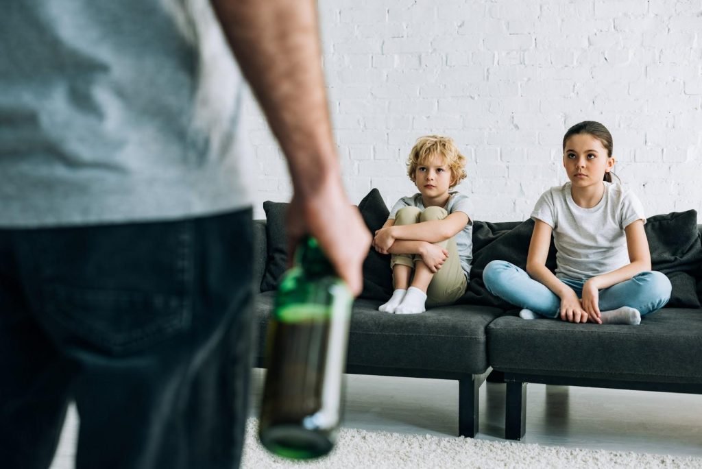 Cropped view of father holding bottle of liquor, facing two small children sitting on sofa; children in divorces involving substance abuse concept.