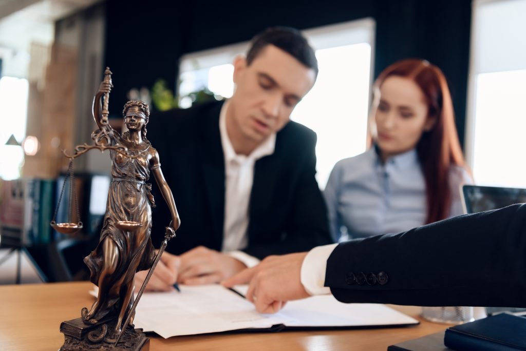 Couple signing documents for divorce or bankruptcy in judge’s office next to desk figurine of blindfolded Justice holding scales