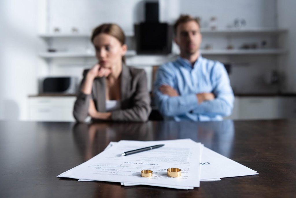 Selective focus of a table with a pair of discarded wedding rings lying atop signed divorce documents in the foreground; divorcing couple seated looking away from each other in the background.