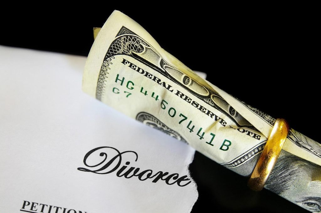 A gold wedding band surrounds a curled hundred dollar bill on top of a ripped paper with the word divorce visible.