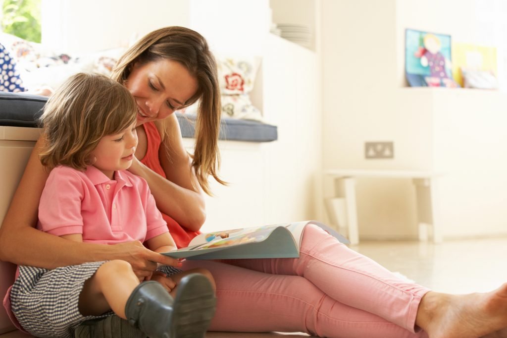 Low-angle view of a young mother sitting on the floor and reading out loud with her arm around a small child; the most common custody schedules by age of child.